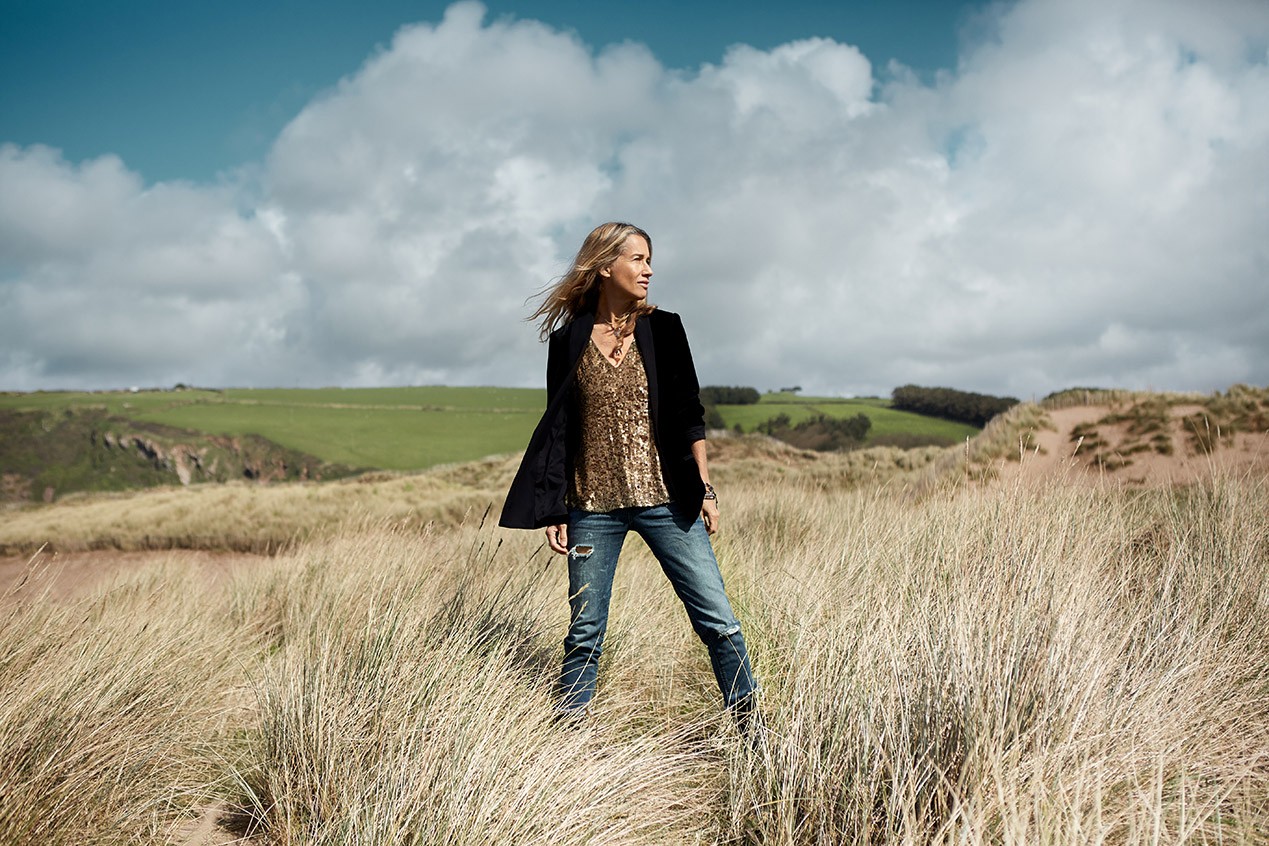 Heather Nova standing in a grassy coastal landscape beneath a cloudy sky, photographed by Louise Roberts.