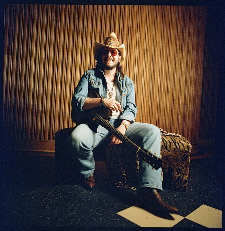 Eddie 9V seated with his guitar, wearing a cowboy hat and denim jacket. Photo by Alysse Gafkjen.