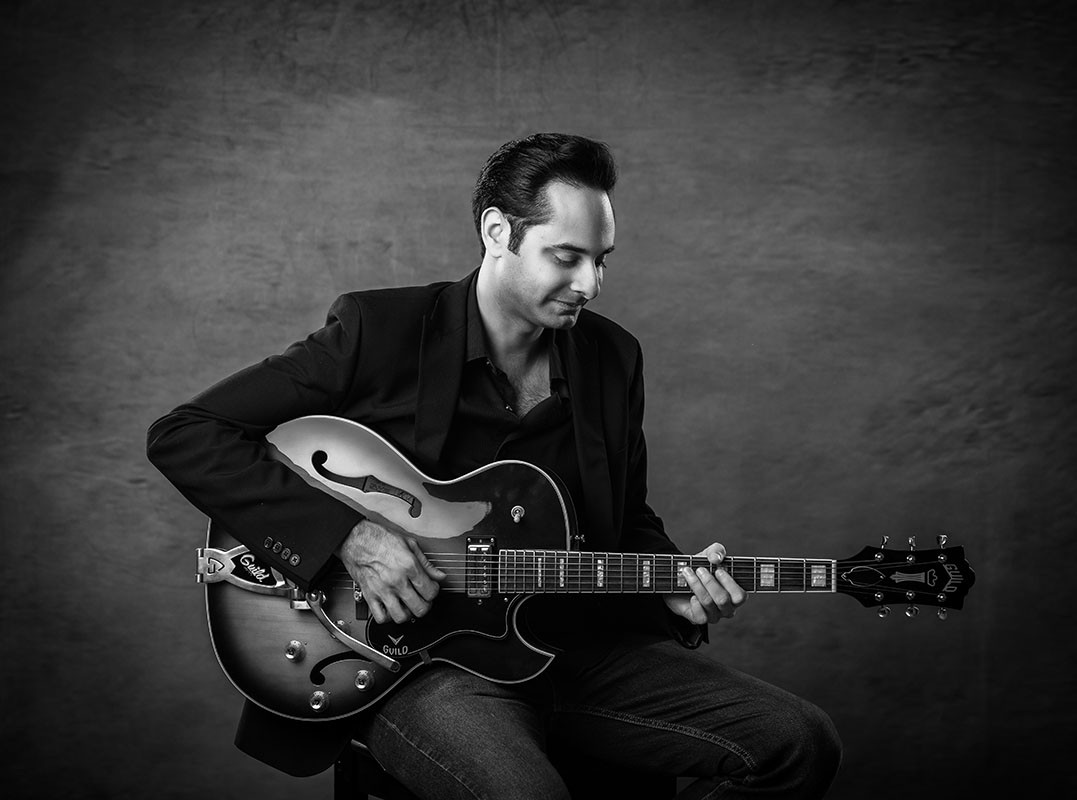 British guitarist Sunjay seated with a Guild electric guitar in a black-and-white promotional photograph 7. Category